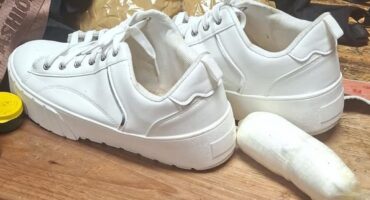 White sneakers on a wooden table with a small package wrapped in plastic and a black backpack partly visible, creating a curious, informal scene.