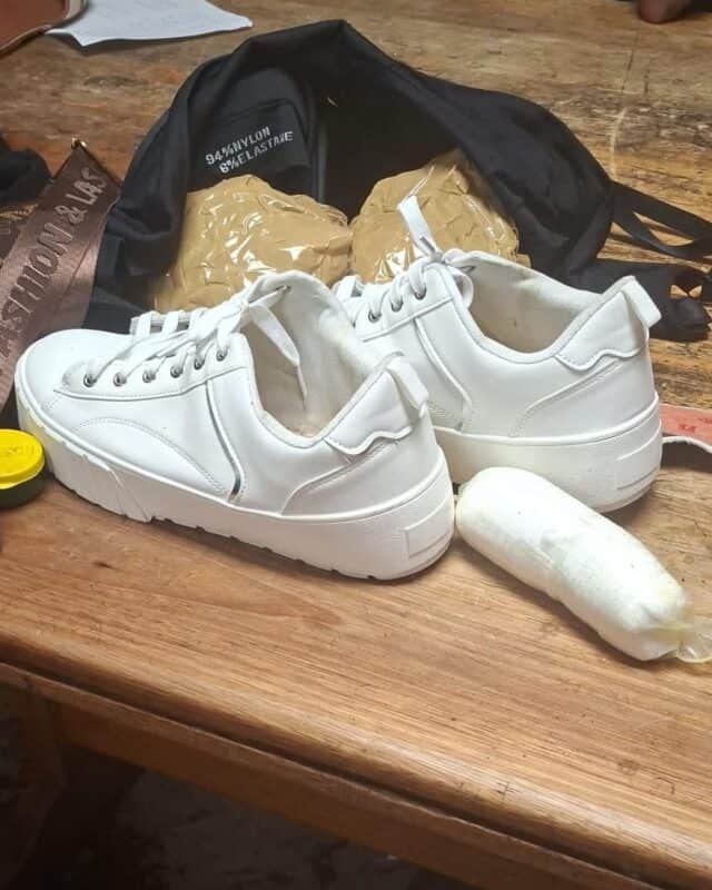 White sneakers on a wooden table with a small package wrapped in plastic and a black backpack partly visible, creating a curious, informal scene.