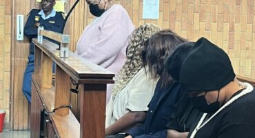 A courtroom scene with four masked individuals sitting in the dock, heads bowed. A police officer stands behind, observing. The mood is tense and solemn.