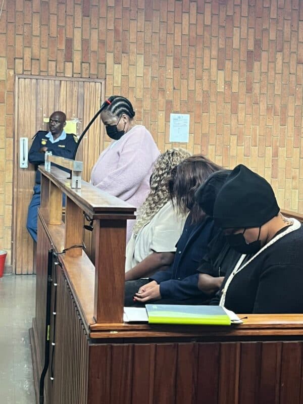 A courtroom scene with four masked individuals sitting in the dock, heads bowed. A police officer stands behind, observing. The mood is tense and solemn.