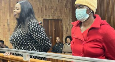 In a wooden-paneled courtroom, two women stand at a podium. One wears a patterned dress, the other a red coat and mask. Observers sit behind them.