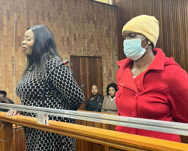 In a wooden-paneled courtroom, two women stand at a podium. One wears a patterned dress, the other a red coat and mask. Observers sit behind them.