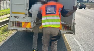 A person in a reflective vest guides another into the back of a large vehicle parked on a highway shoulder under a cloudy sky.