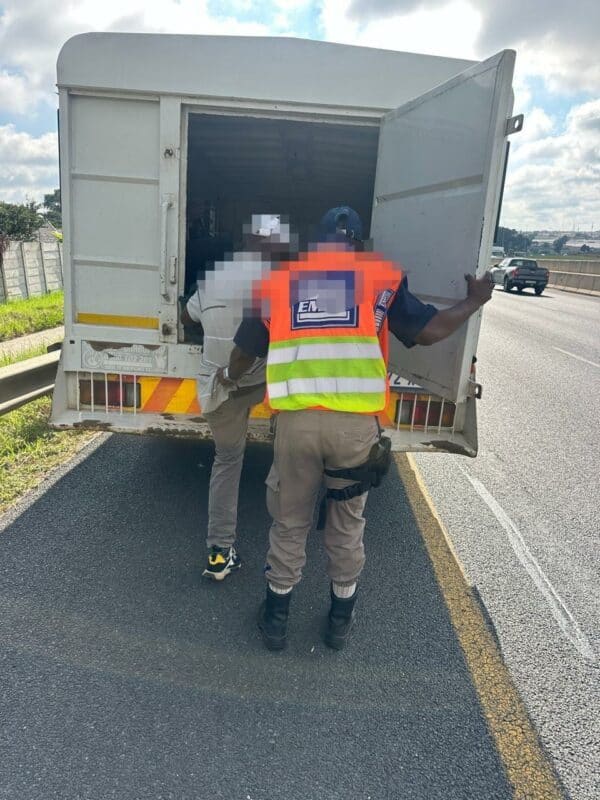 A person in a reflective vest guides another into the back of a large vehicle parked on a highway shoulder under a cloudy sky.