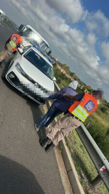 Two people in orange safety vests assist an individual on a highway beside a white car. A tow truck is visible in the background under a cloudy sky.