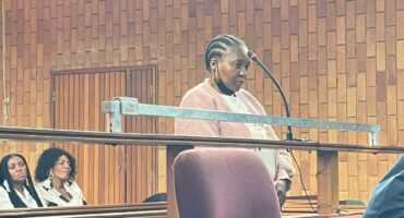 A woman stands at a courtroom witness stand, appearing focused. Behind her, two seated individuals watch attentively. The room features wooden paneling.
