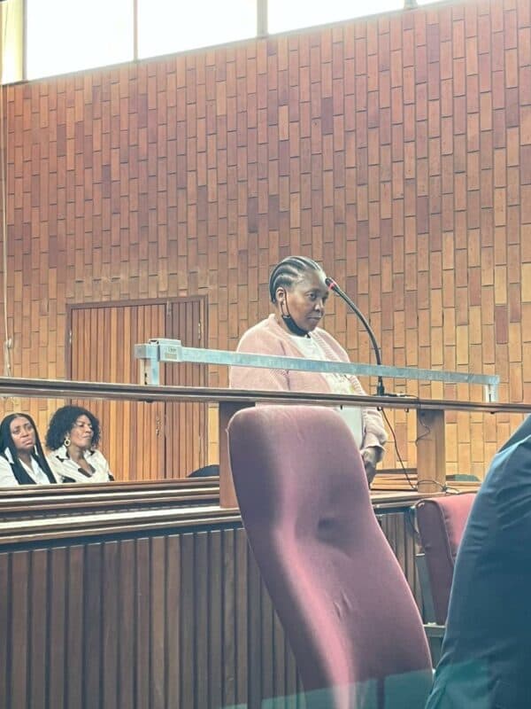 A woman stands at a courtroom witness stand, appearing focused. Behind her, two seated individuals watch attentively. The room features wooden paneling.