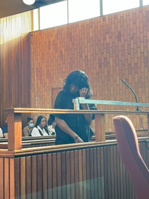 A person stands at a courtroom podium, touching their face, appearing contemplative. Behind them, a brick wall and seated individuals, some wearing masks.