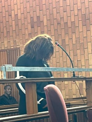 A woman in a black sweater stands in a courtroom, head bowed, holding a microphone. The setting conveys a solemn atmosphere with wooden panels.