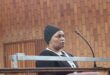 A person stands at a podium with a microphone in a courtroom. The individual wears a black beanie and sweater, appearing serious against a wooden wall backdrop.