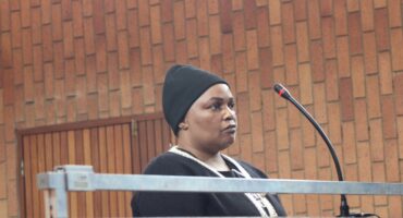 A person stands at a podium with a microphone in a courtroom. The individual wears a black beanie and sweater, appearing serious against a wooden wall backdrop.