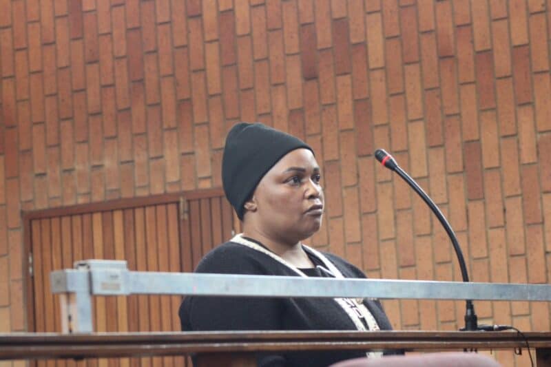 A person stands at a podium with a microphone in a courtroom. The individual wears a black beanie and sweater, appearing serious against a wooden wall backdrop.