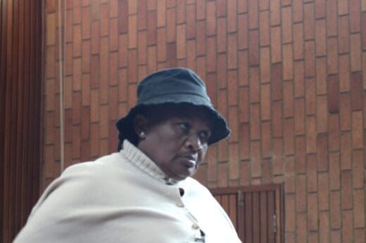 A person wearing a black bucket hat and beige coat stands in front of a brick wall, looking to the side with a serious expression.