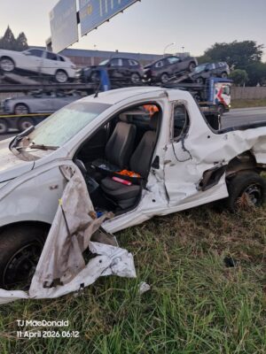 A white car with severe side damage is parked on grass next to a road. It's dawn, and a vehicle transporter carrying cars passes by. The scene conveys a sense of an accident aftermath.