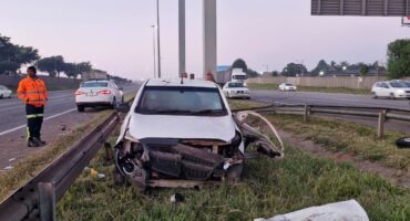 A damaged white car on a grassy roadside with a detached bumper. A person in an orange jacket stands nearby. The sky is overcast with light traffic.