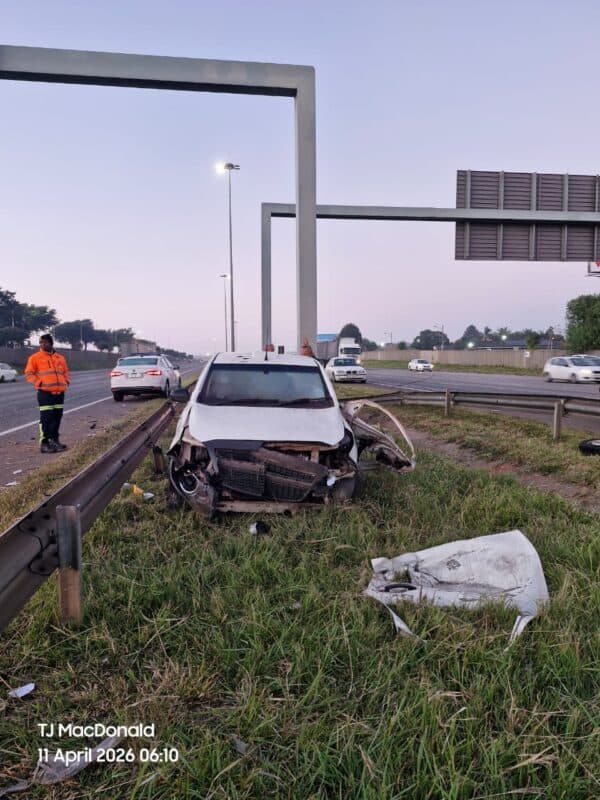 A damaged white car on a grassy roadside with a detached bumper. A person in an orange jacket stands nearby. The sky is overcast with light traffic.