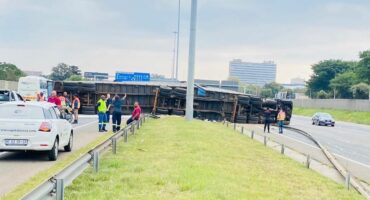 Overturned truck on highway, with people in safety vests assessing the scene. Nearby, a white car is parked, and the sky is cloudy.