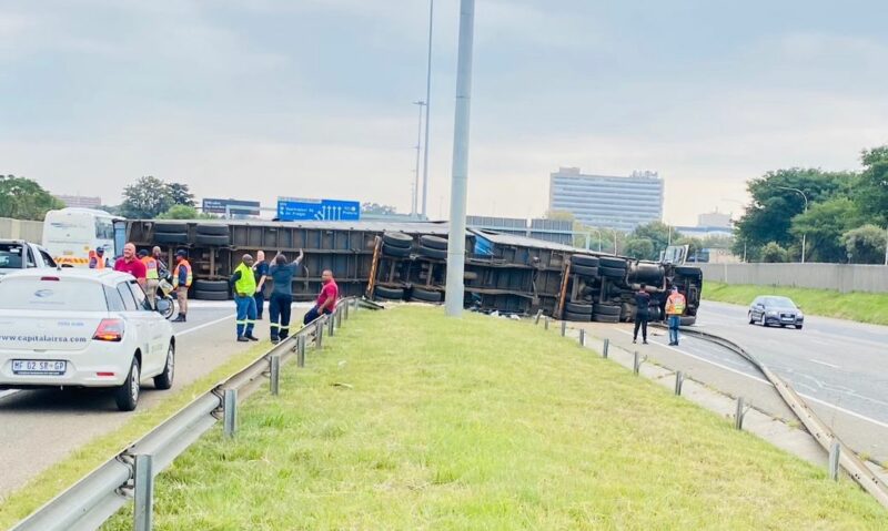 Overturned truck on highway, with people in safety vests assessing the scene. Nearby, a white car is parked, and the sky is cloudy.