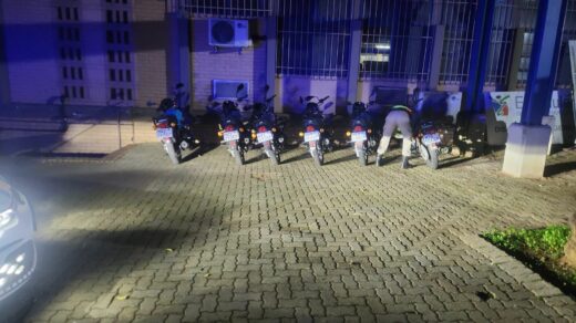 A nighttime scene shows seven motorcycles parked side by side on a lit brick pavement. A person is inspecting one of the motorcycles. The background is an illuminated building with barred windows, creating a secure atmosphere.
