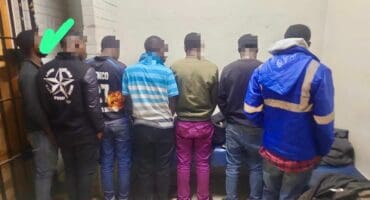 Seven individuals stand with their backs to the camera in a room with white walls. The mood is somber. A green check mark is above one person.