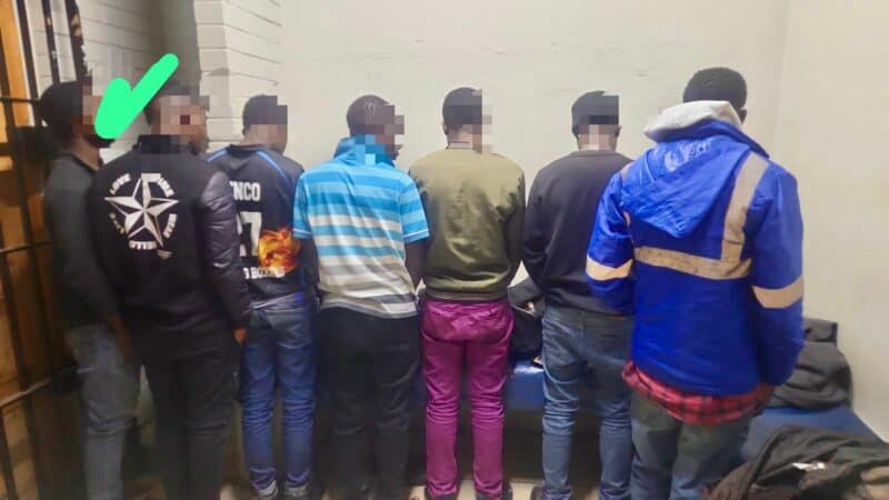 Seven individuals stand with their backs to the camera in a room with white walls. The mood is somber. A green check mark is above one person.