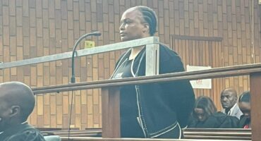 A person standing in a wood-paneled courtroom dock, facing forward, hands behind their back. The mood is tense, with observers seated nearby.