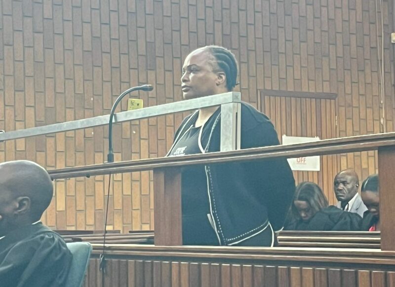 A person standing in a wood-paneled courtroom dock, facing forward, hands behind their back. The mood is tense, with observers seated nearby.