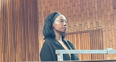 A woman stands in a wood-paneled courtroom, appearing focused and calm. Seated behind her, people watch intently, creating a serious atmosphere.