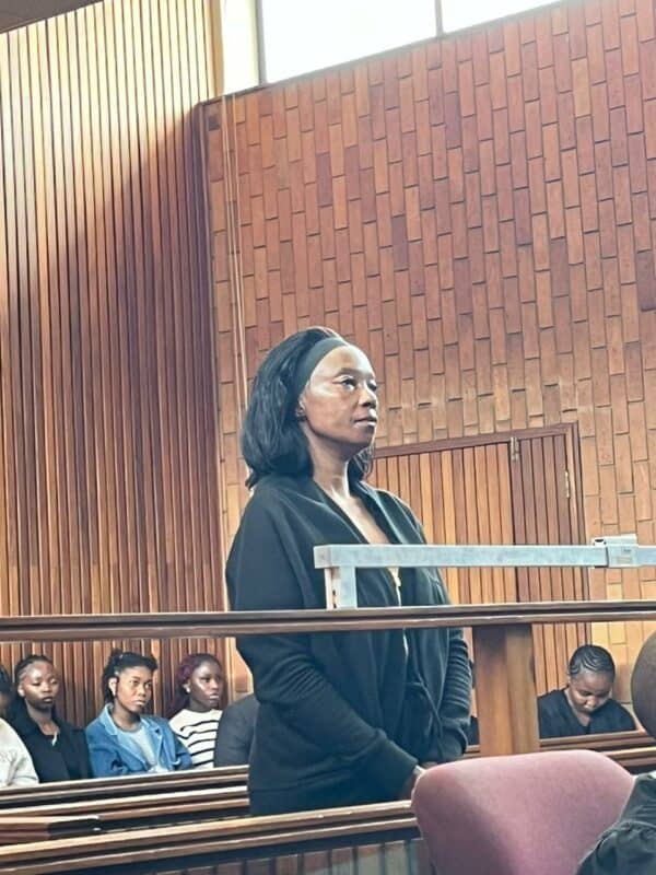 A woman stands in a wood-paneled courtroom, appearing focused and calm. Seated behind her, people watch intently, creating a serious atmosphere.
