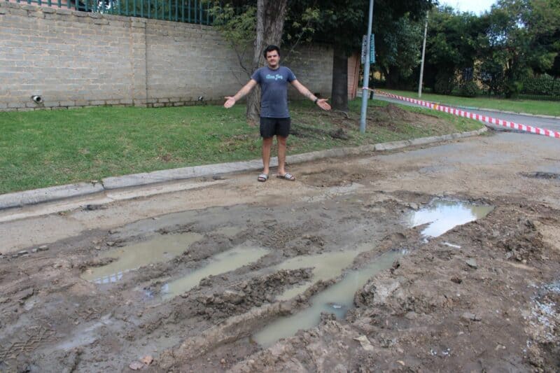 A person stands on a muddy road with large puddles, gesturing in frustration. The street is bordered by a brick wall and a grassy area. The mood is exasperated.