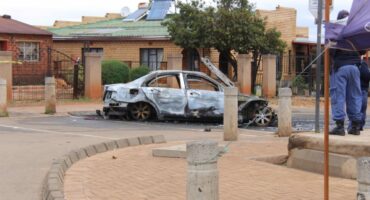Burned-out car on a city street, with charred remains against a residential background. A police officer stands nearby, adding a somber, investigative tone.