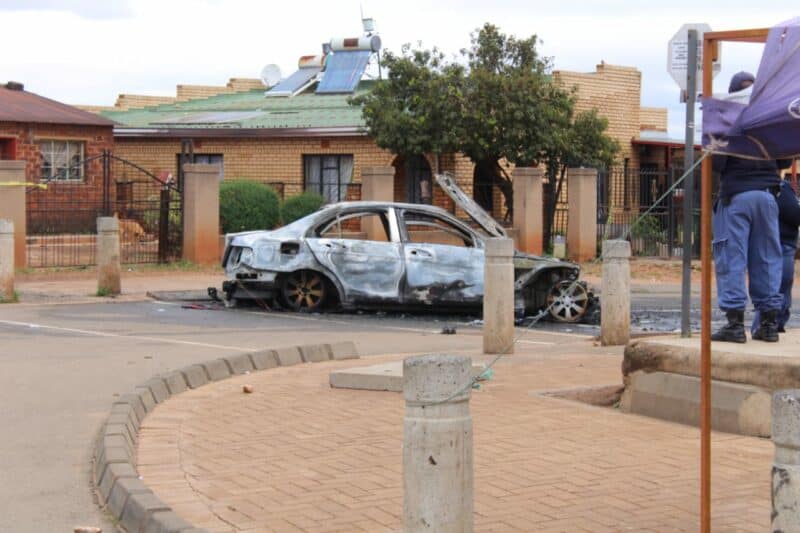Burned-out car on a city street, with charred remains against a residential background. A police officer stands nearby, adding a somber, investigative tone.