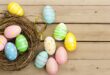 Colorful Easter eggs with various patterns, including stripes and polka dots, are arranged on a wooden surface next to an empty nest. The scene conveys a festive, springtime feel.