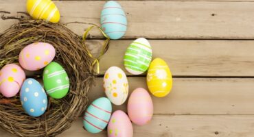 Colorful Easter eggs with various patterns, including stripes and polka dots, are arranged on a wooden surface next to an empty nest. The scene conveys a festive, springtime feel.