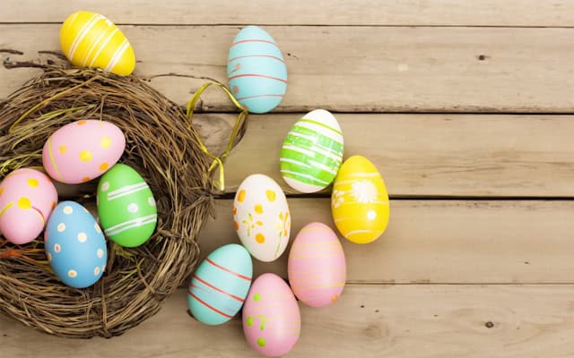 Colorful Easter eggs with various patterns, including stripes and polka dots, are arranged on a wooden surface next to an empty nest. The scene conveys a festive, springtime feel.