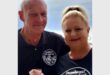 A smiling man and woman stand together, wearing matching black shirts with "December 2022 KZN South Coast" text. The woman has her hair tied up, and the man holds a drink. The background is a soft blue, hinting at a coastal setting, conveying a warm, celebratory vibe.
