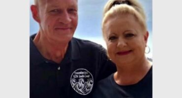 A smiling man and woman stand together, wearing matching black shirts with "December 2022 KZN South Coast" text. The woman has her hair tied up, and the man holds a drink. The background is a soft blue, hinting at a coastal setting, conveying a warm, celebratory vibe.