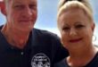 A smiling man and woman stand together, wearing matching black shirts with "December 2022 KZN South Coast" text. The woman has her hair tied up, and the man holds a drink. The background is a soft blue, hinting at a coastal setting, conveying a warm, celebratory vibe.