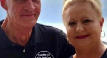 A smiling man and woman stand together, wearing matching black shirts with "December 2022 KZN South Coast" text. The woman has her hair tied up, and the man holds a drink. The background is a soft blue, hinting at a coastal setting, conveying a warm, celebratory vibe.