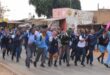 A group of energetic students in uniforms run down a street lined with shops. Their expressions are joyful and excited, conveying a lively atmosphere.