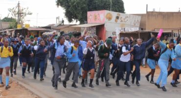 A group of energetic students in uniforms run down a street lined with shops. Their expressions are joyful and excited, conveying a lively atmosphere.