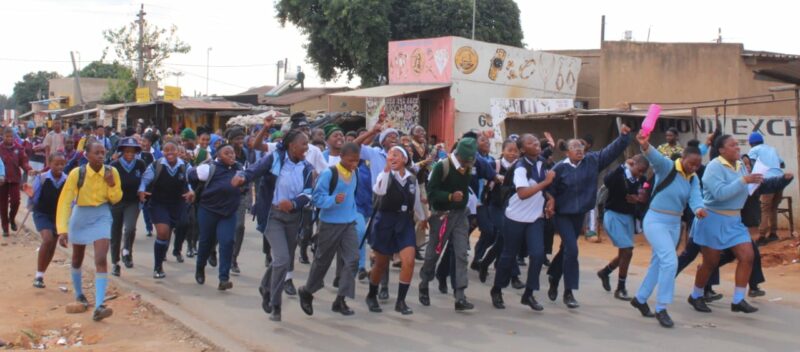 A group of energetic students in uniforms run down a street lined with shops. Their expressions are joyful and excited, conveying a lively atmosphere.