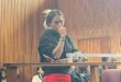 A woman in a black top and red skirt stands pensively in a wood-paneled courtroom. Seated people in the background appear attentive. The atmosphere is tense and serious.