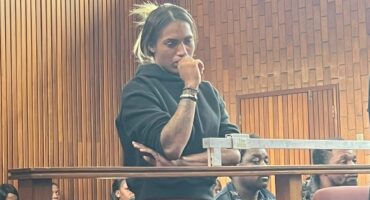 A woman in a black top and red skirt stands pensively in a wood-paneled courtroom. Seated people in the background appear attentive. The atmosphere is tense and serious.
