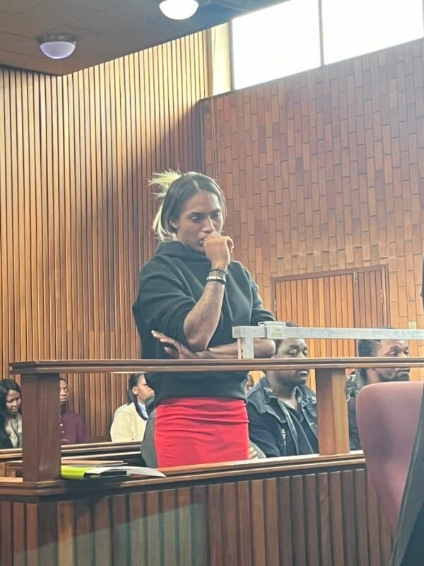 A woman in a black top and red skirt stands pensively in a wood-paneled courtroom. Seated people in the background appear attentive. The atmosphere is tense and serious.