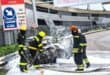 Firefighters in protective gear extinguish a car fire with foam on a city street. Surroundings include a bus sign and large billboard. Dramatic scene.