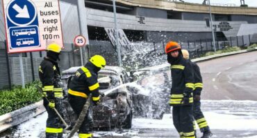 Firefighters in protective gear extinguish a car fire with foam on a city street. Surroundings include a bus sign and large billboard. Dramatic scene.