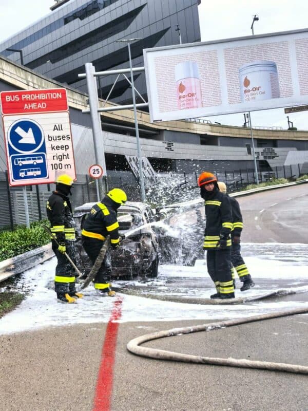Firefighters in protective gear extinguish a car fire with foam on a city street. Surroundings include a bus sign and large billboard. Dramatic scene.