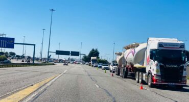Traffic scene on a highway under a clear blue sky, with a line of trucks and cars stopped to the right, and a sign showing directions to Pretoria.