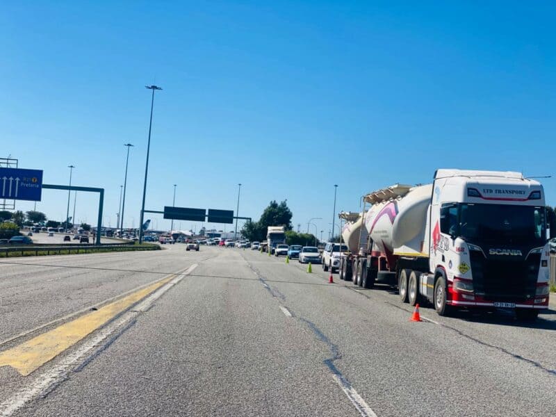 Traffic scene on a highway under a clear blue sky, with a line of trucks and cars stopped to the right, and a sign showing directions to Pretoria.
